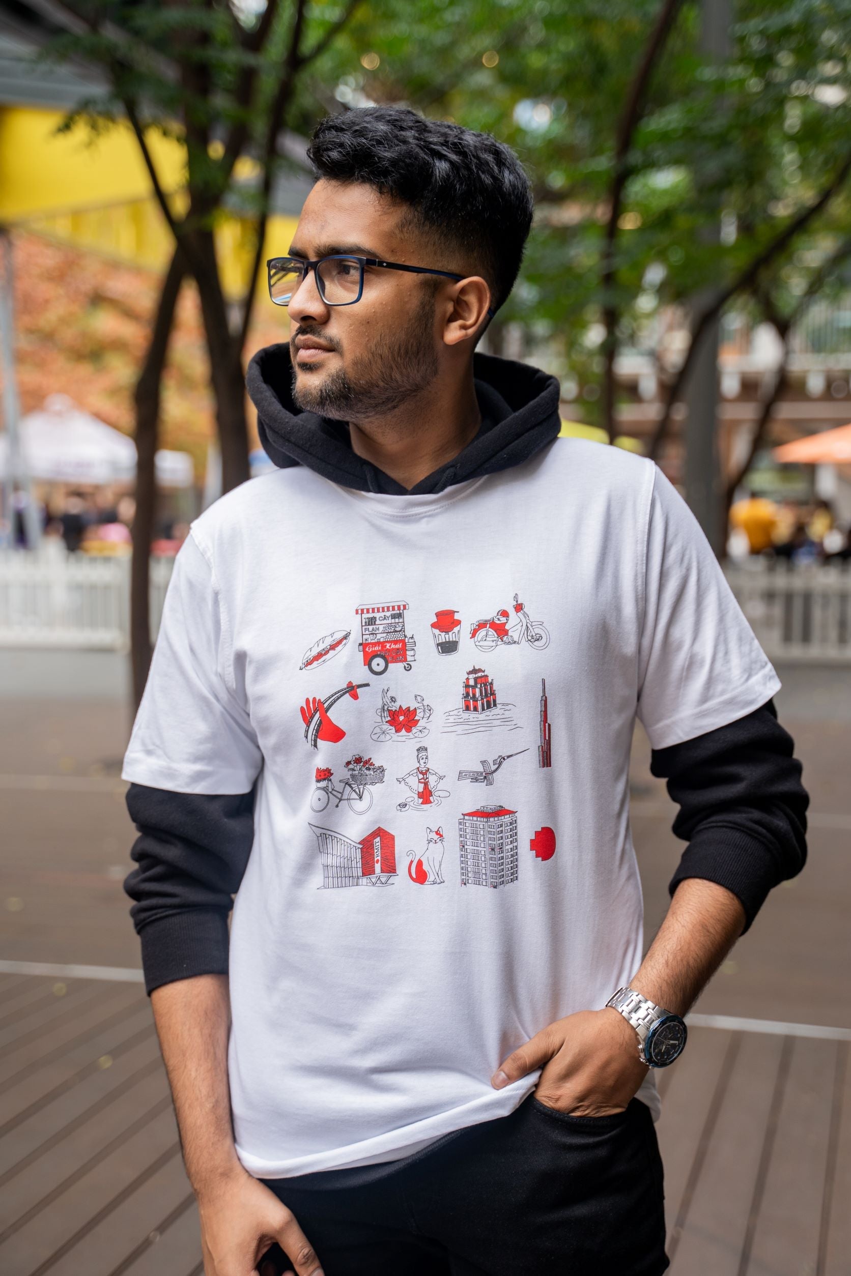 Male model displays the front of the Vietnamese Icon Tee from RMIT Store. The T-shirt features vibrant red and black illustrations of Vietnamese cultural heritage against the white fabric. He pairs the tee with black pants, standing in a leafy outdoor setting.